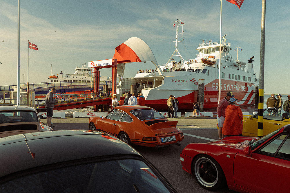 Ein orangefarbener Porsche 911 wartet im Vordergrund auf die Einfahrt in die Fähre, im Hintergrund das FRS Syltfähre Schiff SyltExpress am Anleger in Havneby.