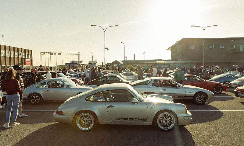 Porsche Klassiker verschiedener Baujahre stehen im Abendlicht auf dem Parkplatz am Fährhafen Havneby, im Vordergrund ein silberner Porsche 964 mit seitlichem Porsche-Schriftzug.