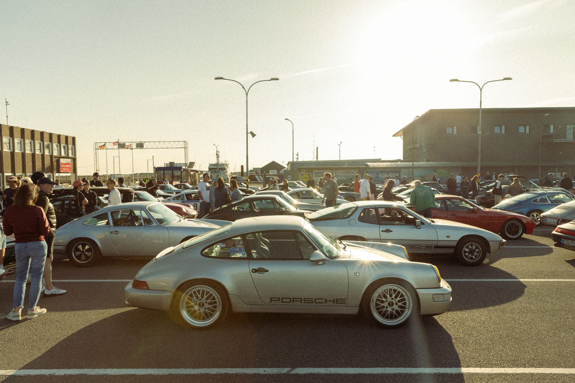 Porsche Klassiker verschiedener Baujahre stehen im Abendlicht auf dem Parkplatz am Fährhafen Havneby, im Vordergrund ein silberner Porsche 964 mit seitlichem Porsche-Schriftzug.